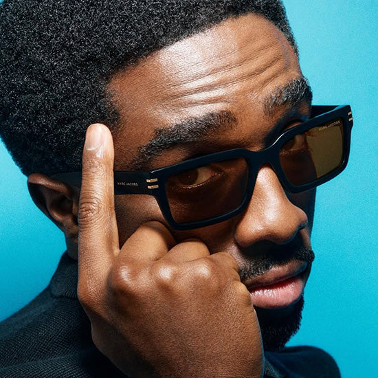 Ritratto ravvicinato di un uomo con occhiali da sole Marc Jacobs dalla montatura squadrata nera e dettagli raffinati sulle aste. L'immagine mette in evidenza il design moderno, l'eleganza urbana e l'estetica premium tipica dell'eyewear di lusso.
