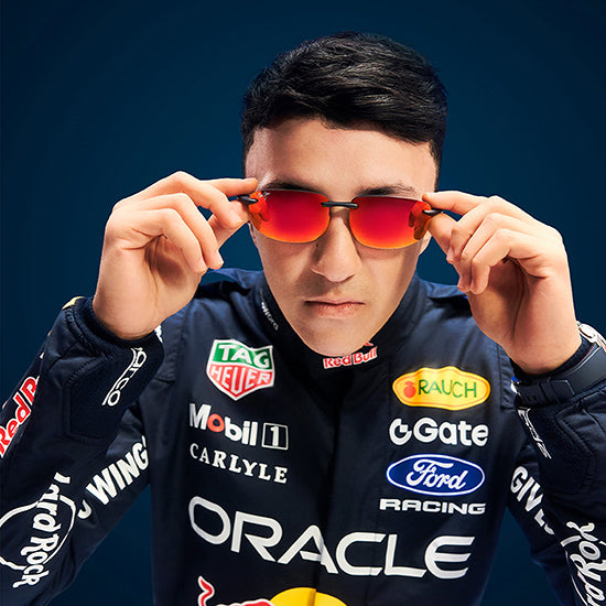 Pilota del team Oracle Red Bull Racing indossa occhiali da sole sportivi Maui Jim con lenti specchiate rosso-arancio, mentre regola la montatura guardando verso l'obiettivo su sfondo blu scuro.
