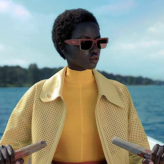 Donna con occhiali da sole Salvatore Ferragamo caratterizzati da una montatura geometrica moderna in tonalità terracotta. L'ambientazione sul lago e l'abbigliamento dai colori vivaci richiamano eleganza contemporanea, lusso e stile estivo raffinato.